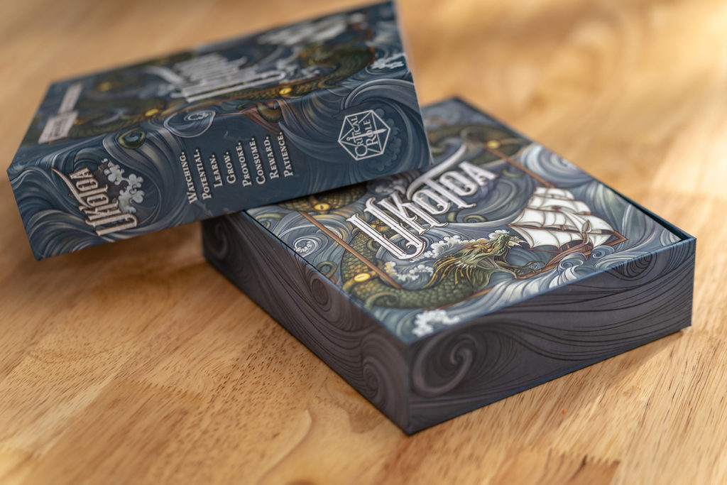 A photograph of the Uk'otoa box on a table, with the cover taken off and propped at a jaunty angle, revealing the rulebook on top of the contents inside.