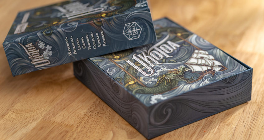 A photograph of the Uk'otoa box on a table, with the cover taken off and propped at a jaunty angle, revealing the rulebook on top of the contents inside.