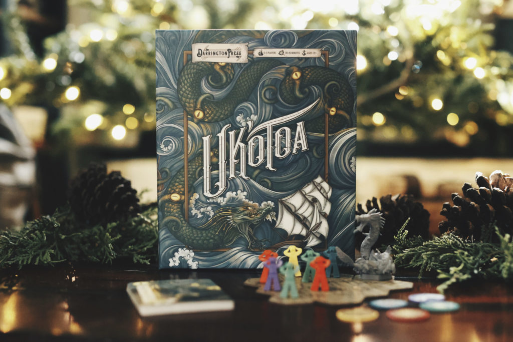 A photo of Uk'otoa, the board game, with meeples, tiles, miniature, and cards in the foreground and lit-up trees in the background.