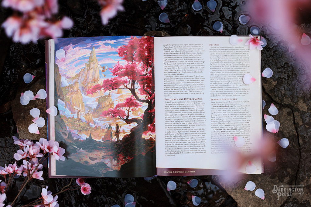 A photo of the book featuring flower blossoms on a stony trickling water backround, open to a page featuring an illustration of the landscape of Zephrah: A village among the clouds on grassy, spiralling mountain peaks. There is a tall, purple leafed tree in the foreground with the druid Keyleth and her mother Vilya holding staffs, conversing underneath it as they look at the village. Two individuals on skysails fly overhead.